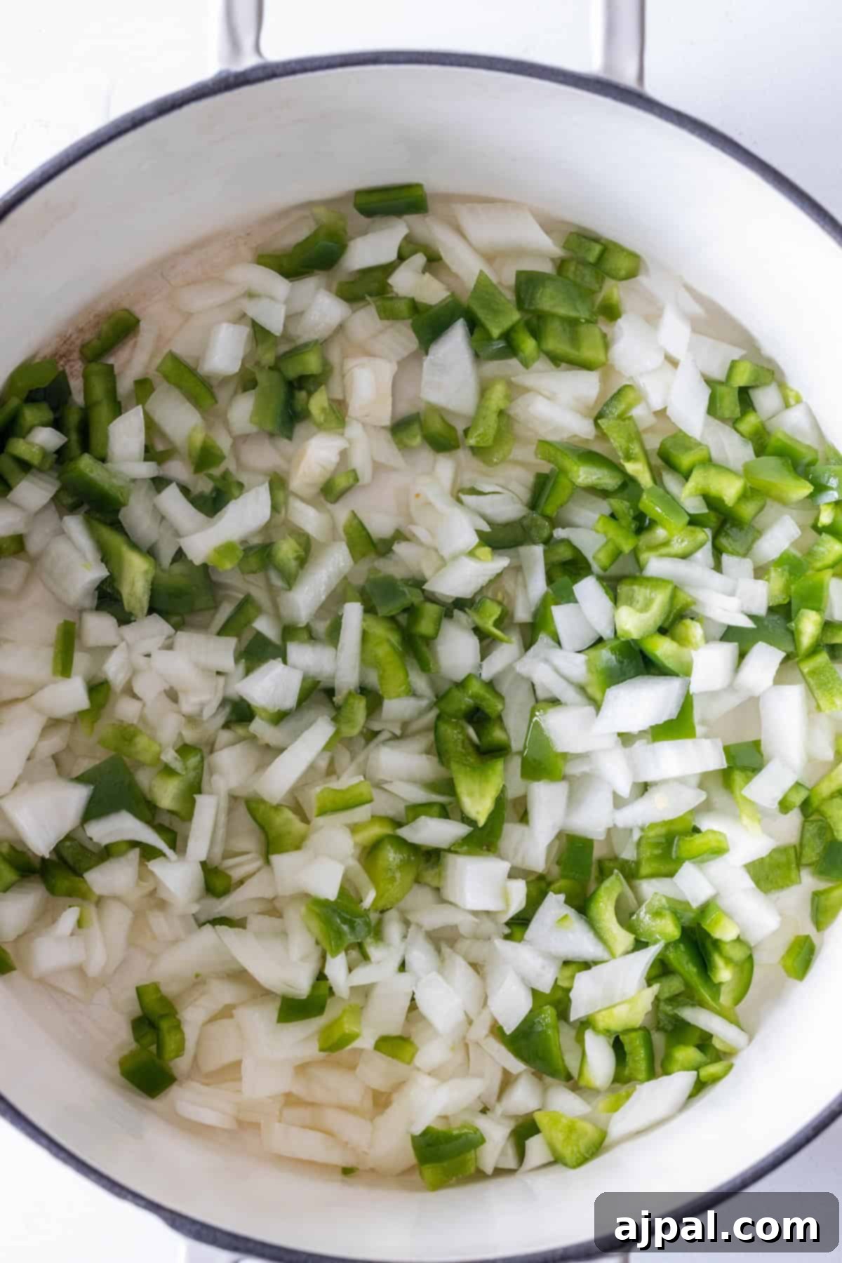 Chopped white onions and green bell peppers in a white pot, ready to be cooked for a flavorful vegan rice soup.