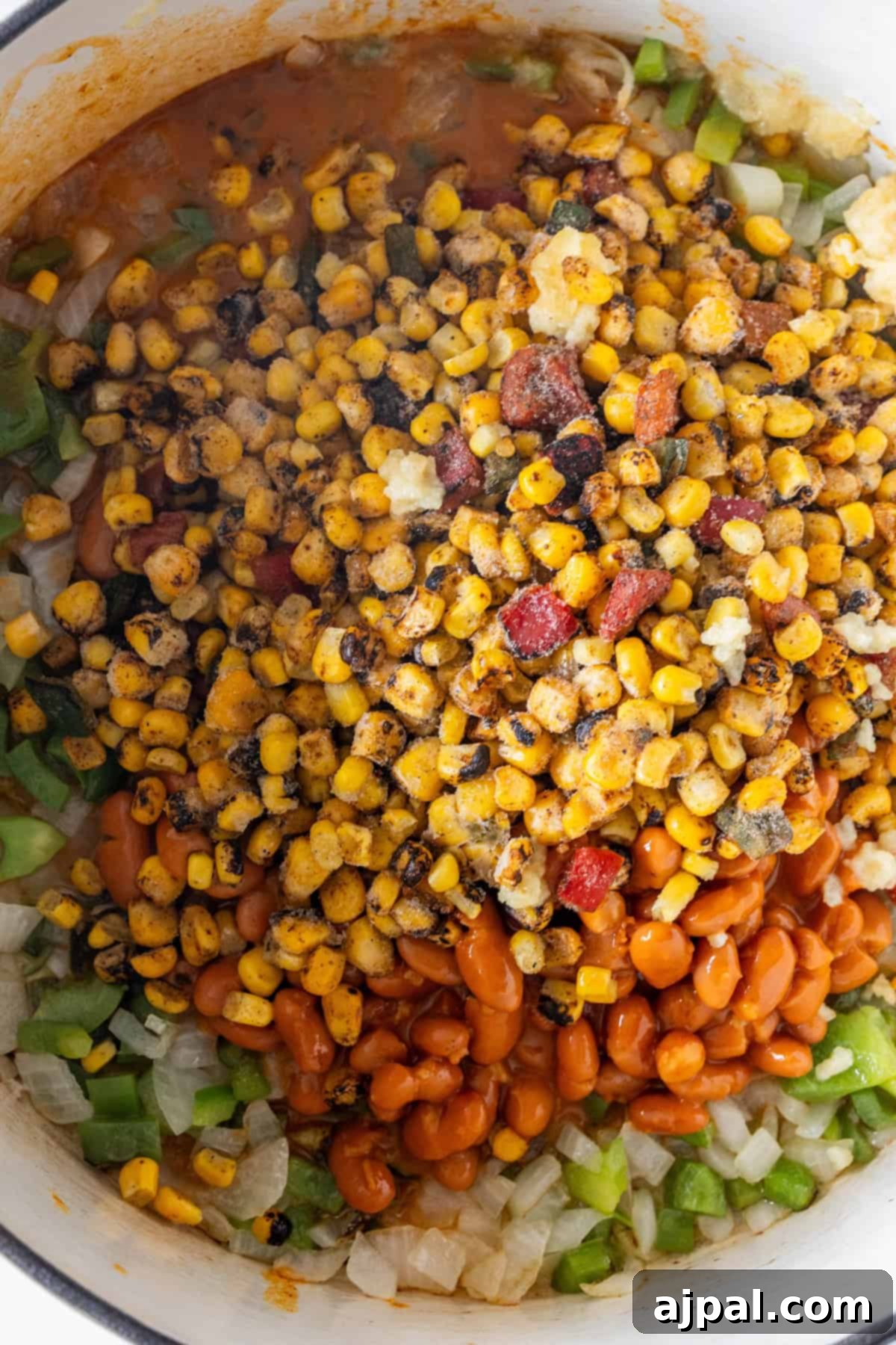 A close-up of a pot containing roasted corn, pinto beans, chopped green bell pepper, and diced onions in a tomato-based sauce—perfect for a hearty Vegan Charro Bean soup.