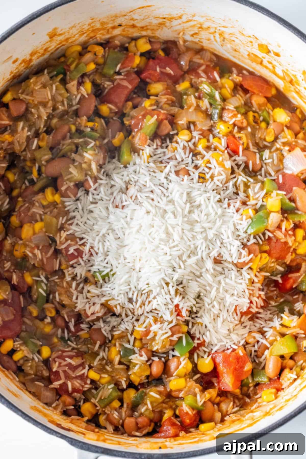 A pot filled with a mixture of charro beans, corn, tomatoes, peppers, and uncooked white rice on top, ready to be stirred into a hearty vegan soup.