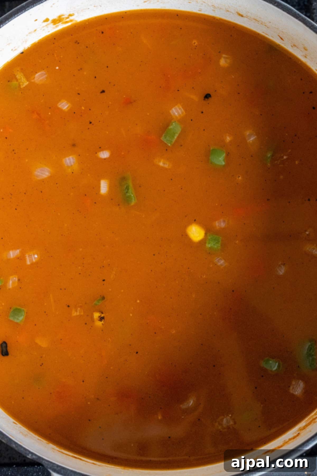 A pot of orange-red vegan Charro Bean soup with visible chopped vegetables such as green beans, corn, and onions.