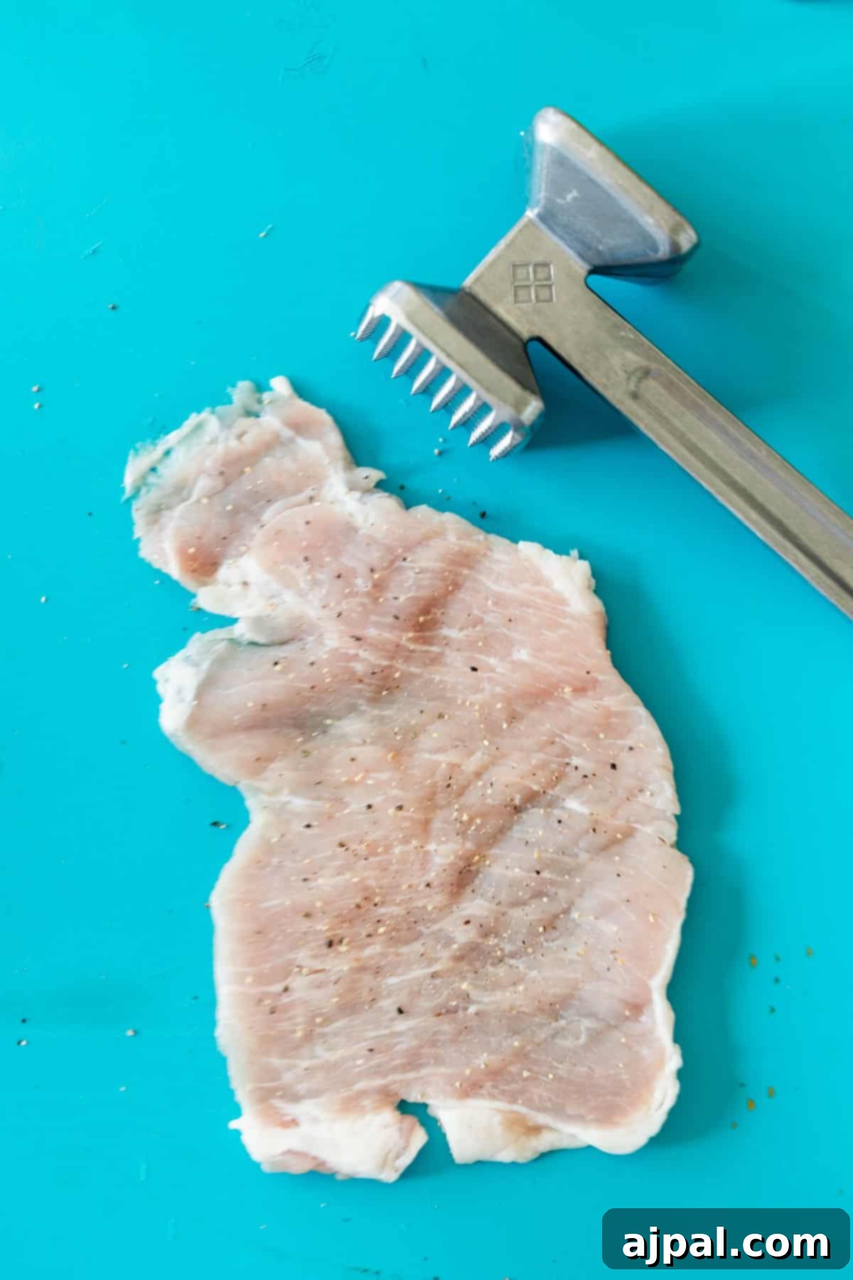 Crispy Oven Pork Cutlets 5 Raw pork chop being pounded thin on a blue cutting board with a meat tenderizer. This step is crucial for even cooking and tenderness in baked breaded pork chops.