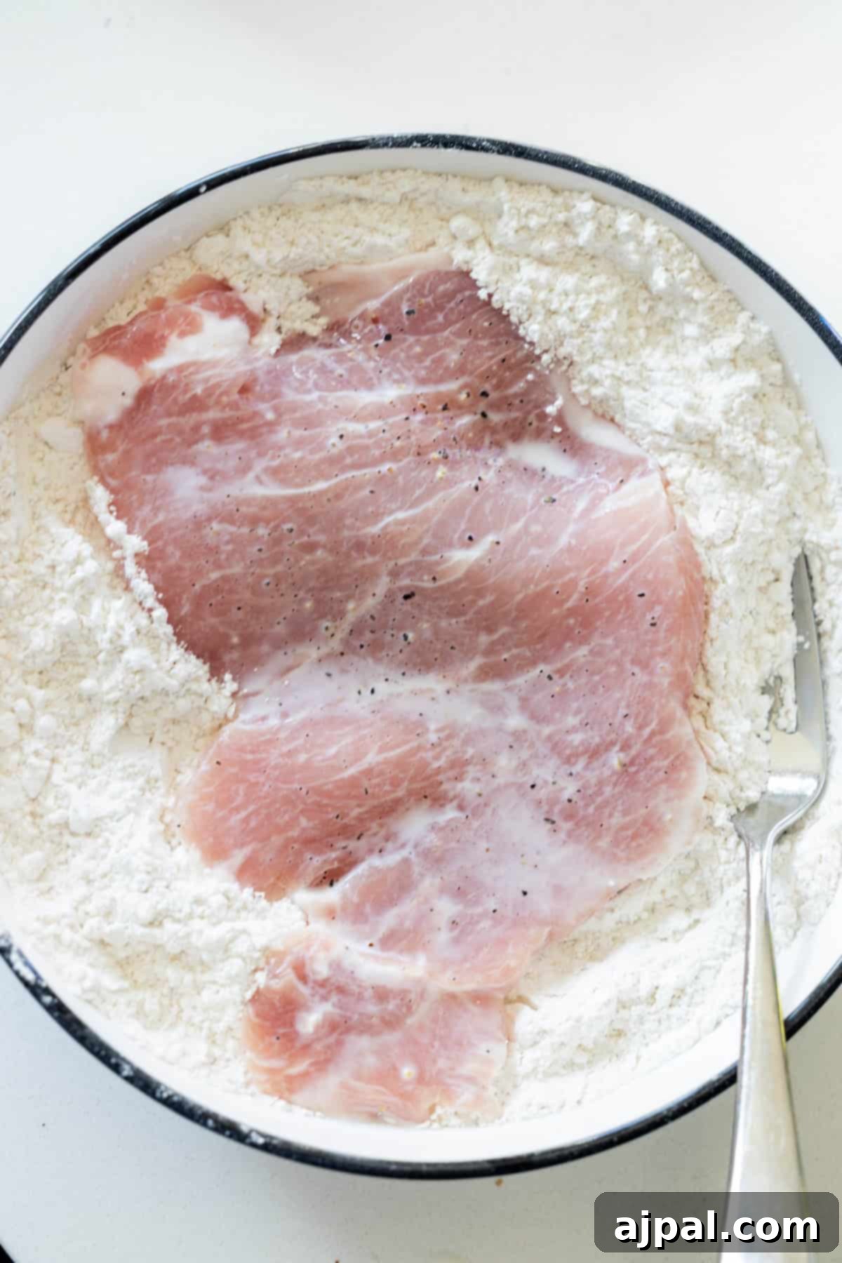 Crispy Oven Pork Cutlets 7 A raw pork chop coated in flour, resting in a shallow white dish, ready for the next step in preparing crispy baked breaded pork chops.