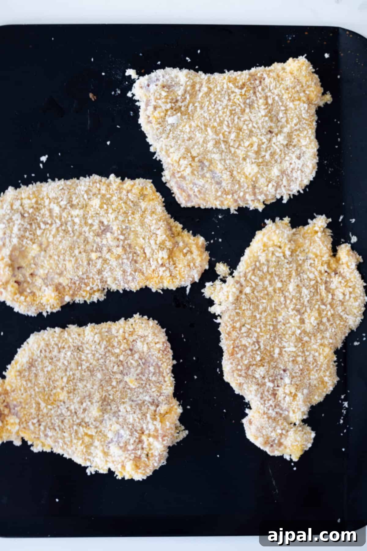 Crispy Oven Pork Cutlets 10 Four raw breaded pork chops arranged neatly on a dark baking surface, ready for the oven to transform them into delicious, crispy baked breaded pork chops.
