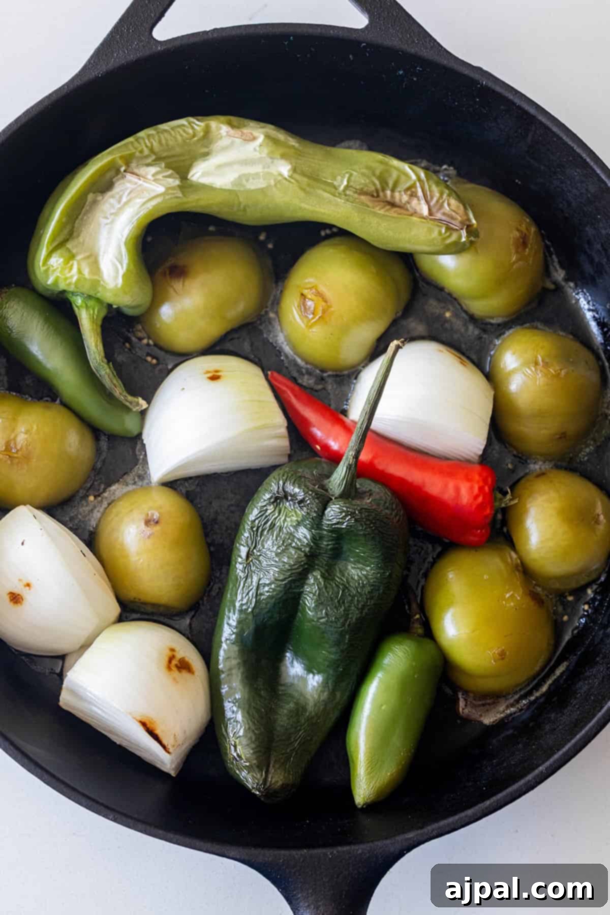 A cast iron skillet with tomatillos, white onion chunks, jalapeño, Anaheim, and red chili peppers being roasted for a flavorful roasted salsa.