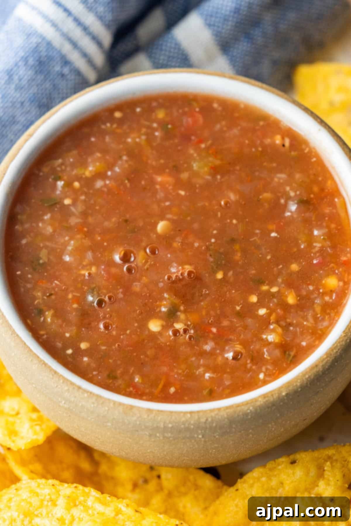 A bowl of chunky roasted salsa sits next to yellow tortilla chips with a blue and white cloth in the background.