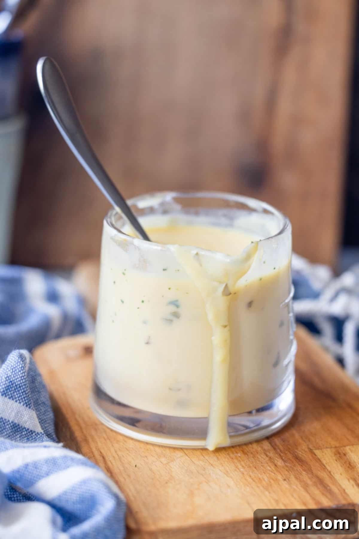 Greek Lemon-Egg Sauce 3 A glass filled with creamy Avgolemono Sauce, some dripping down the side, with a spoon inside, placed on a wooden board next to a blue and white cloth.