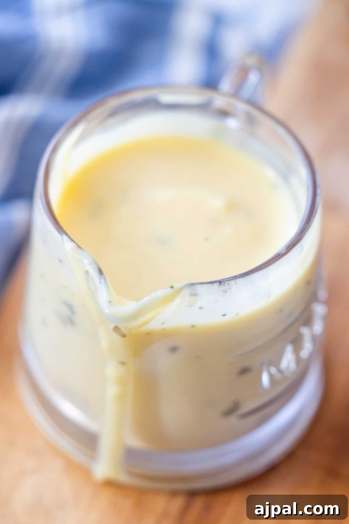 Greek Lemon-Egg Sauce 4 A clear glass measuring cup filled with creamy yellow Greek Avgolemono Sauce, with some sauce dripping down the side, sits on a wooden surface.