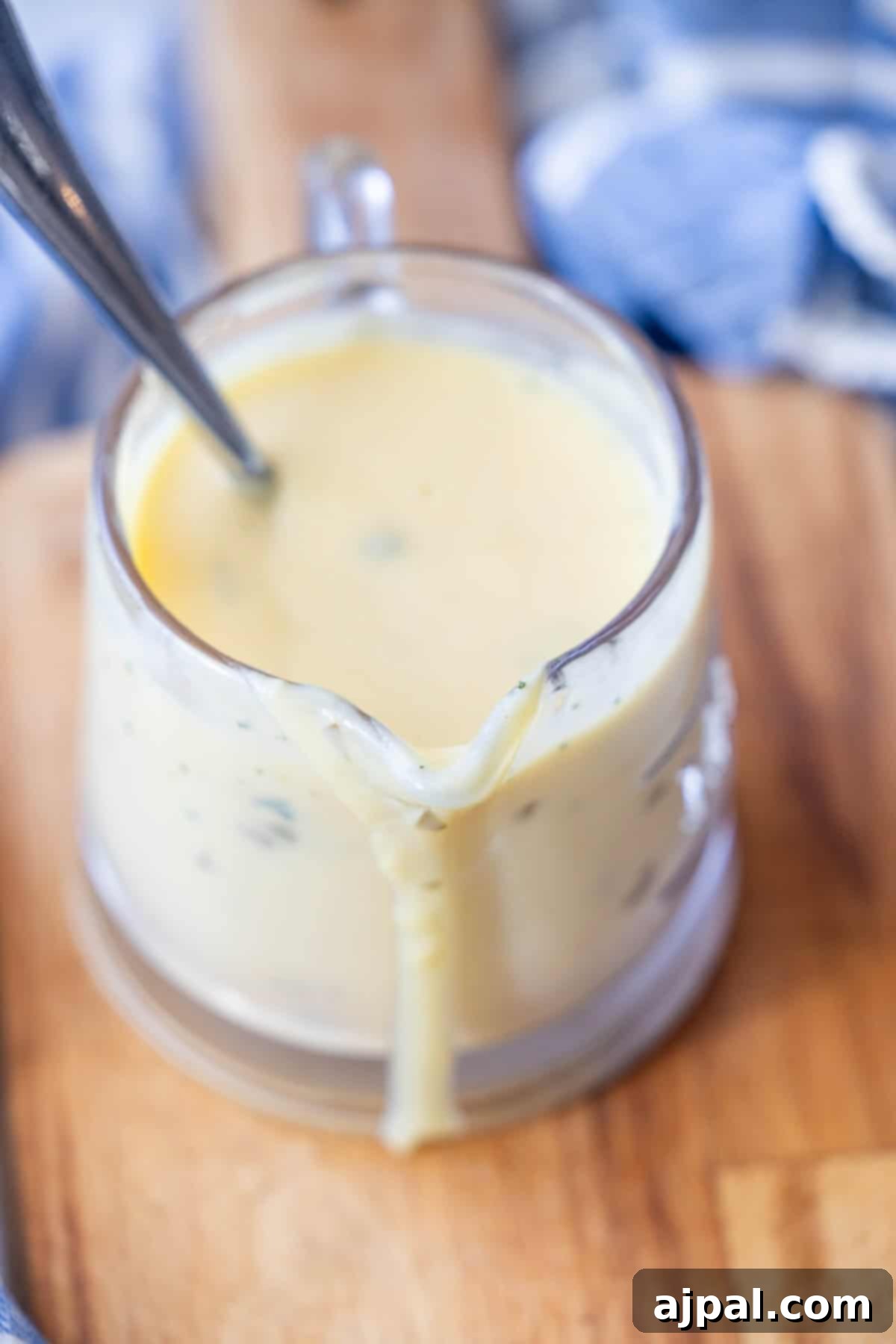 Greek Lemon-Egg Sauce 5 A glass measuring cup filled with creamy yellow avgolemono sauce, with a spoon inside and sauce dripping down the spout, sits on a wooden surface.