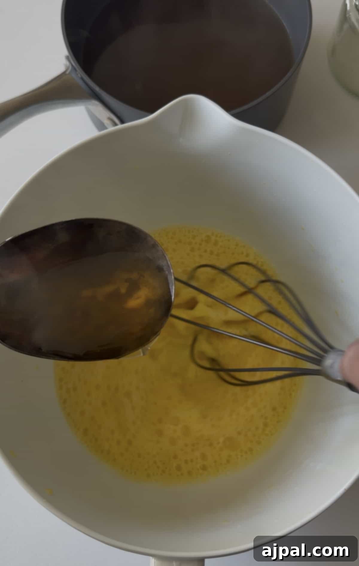 Greek Lemon-Egg Sauce 7 A metal spoon pours liquid into a bowl of beaten eggs being whisked, as Avgolemono sauce—a classic Greek sauce—comes together, with a saucepan and jar in the background.