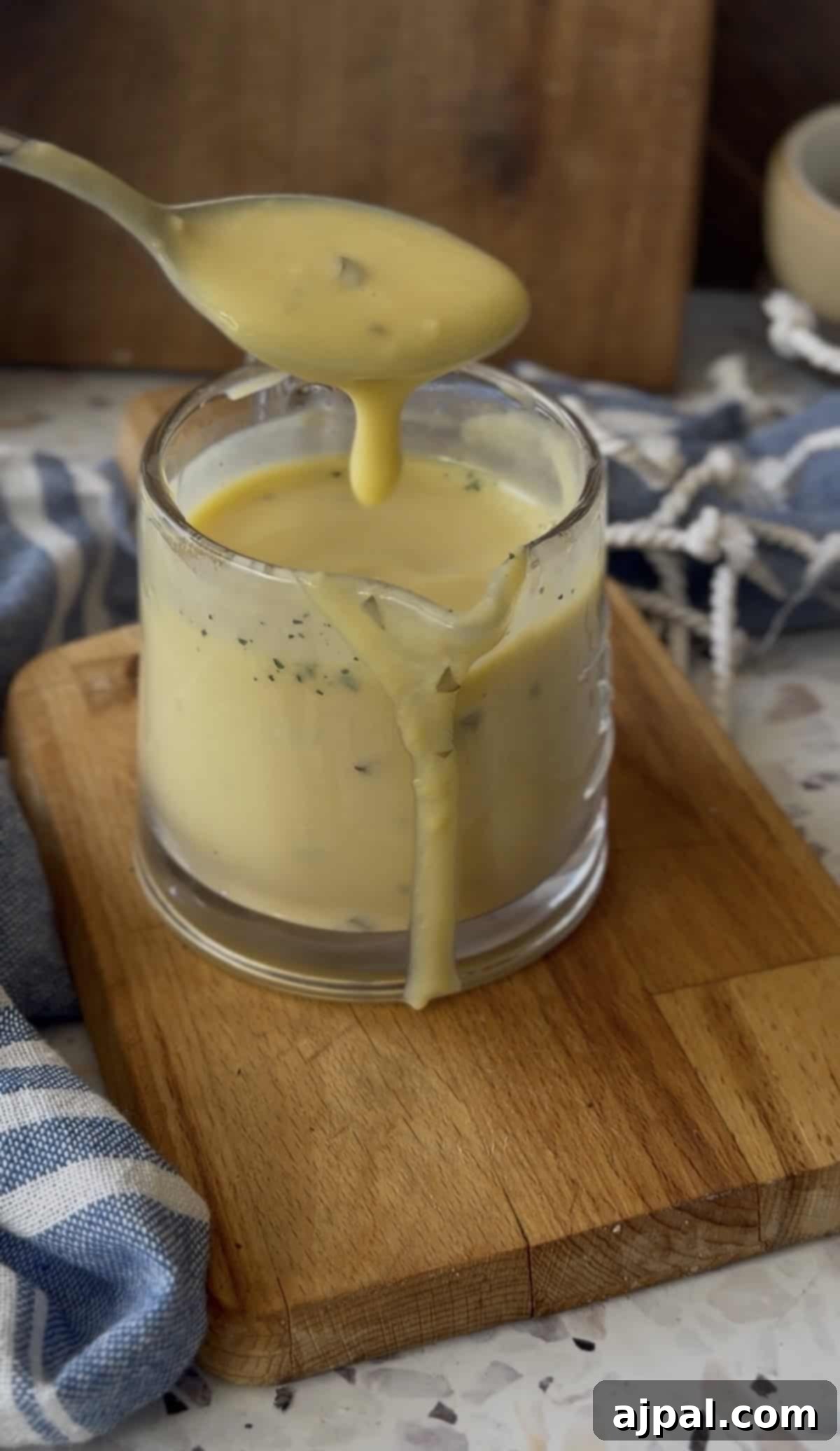 Greek Lemon-Egg Sauce 9 A spoon pours thick yellow Avgolemono Sauce with herbs into a glass cup, causing some sauce to drip down the side onto a wooden board.