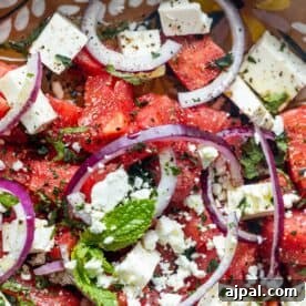 Close-up of a Watermelon Feta Salad with feta cheese cubes, sliced red onions, fresh mint leaves, basil, chopped herbs, and ground black pepper.