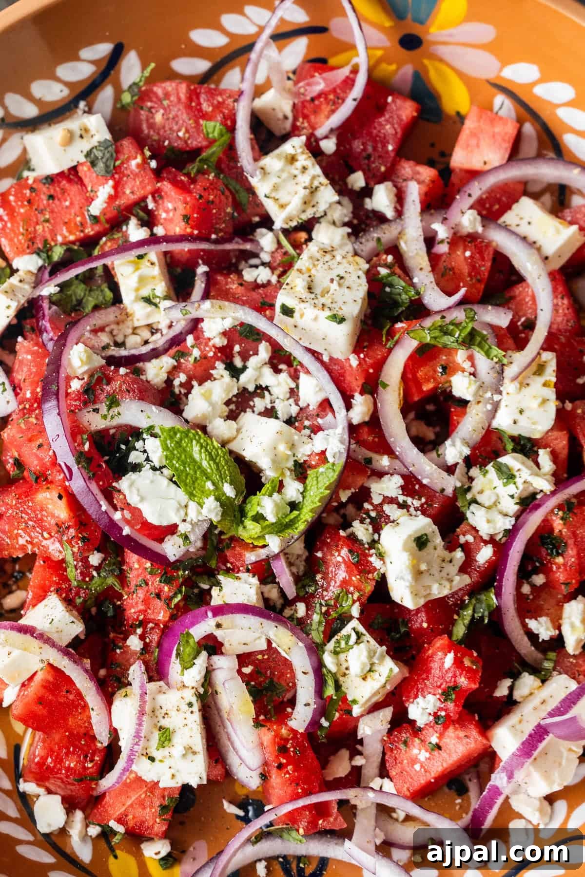 A close-up of a watermelon feta salad with juicy watermelon cubes, creamy feta cheese, red onion slices, fresh mint, basil, black pepper, and herbs on a decorative plate.