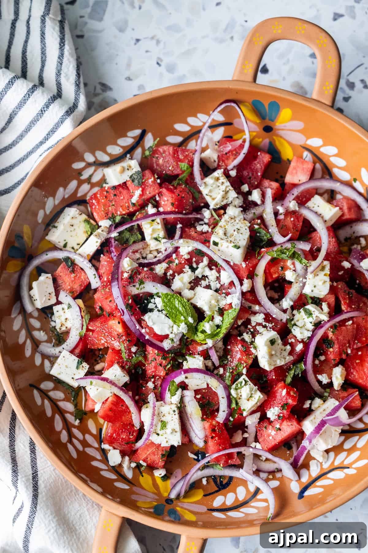 A refreshing Watermelon Feta Salad with cubed watermelon, feta cheese, red onion slices, mint and basil leaves, and seasoning, served in a decorative orange bowl on a countertop.
