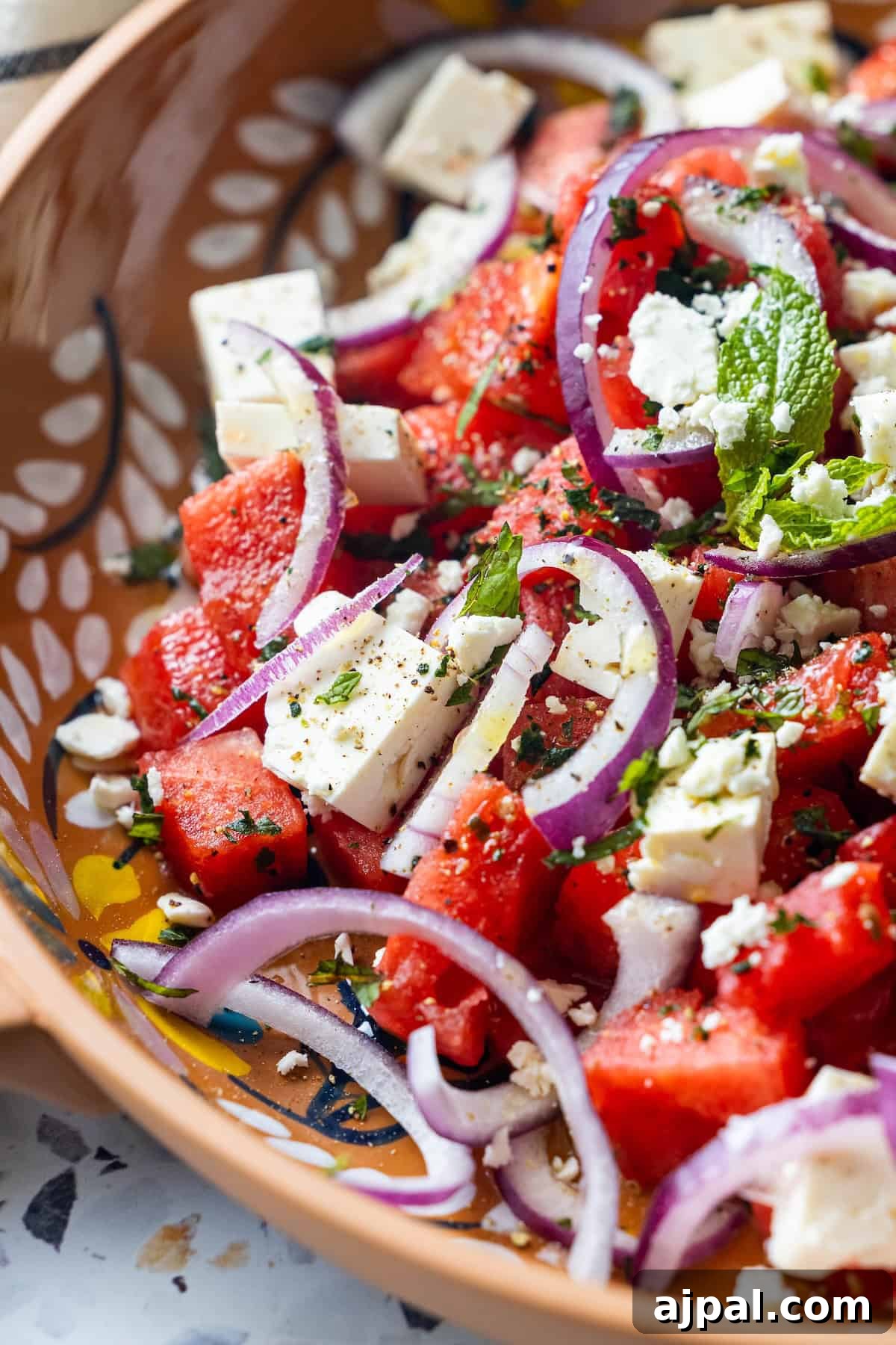 A close-up of a Watermelon Feta Salad with juicy watermelon cubes, feta cheese, red onion slices, fresh mint and basil, finished with black pepper in a decorative ceramic bowl.