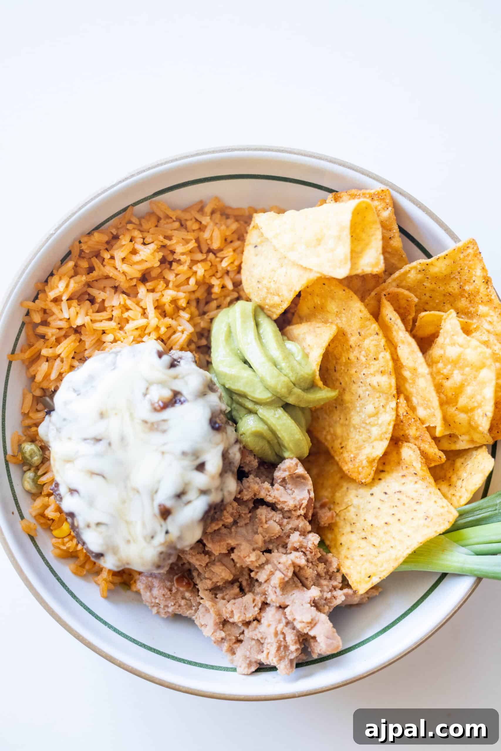A bowl featuring Mexican rice, tortilla chips, refried beans, ground beef with melted cheese and green chile, topped with guacamole and garnished with a green onion