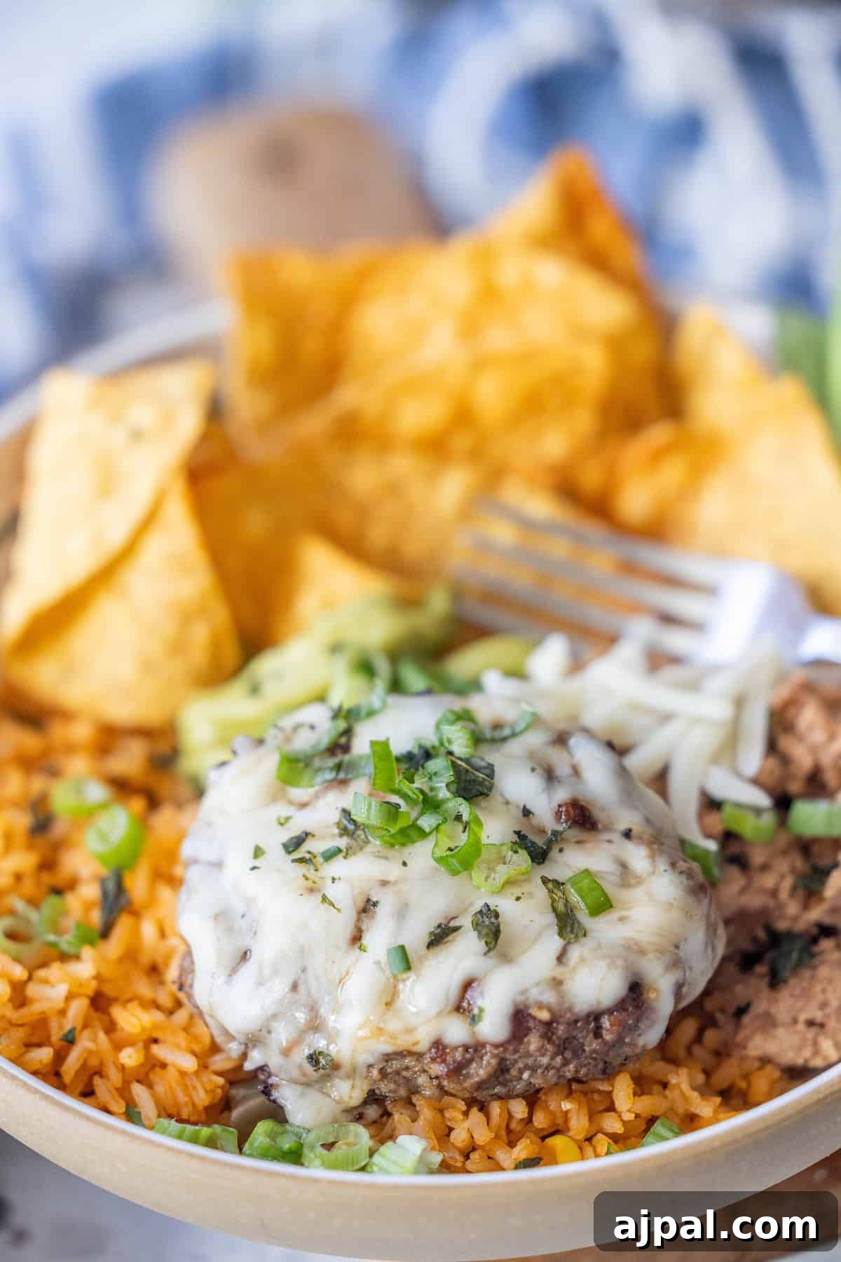 A plate with Mexican rice, a Green Chile Burger patty topped with melted cheese and herbs, avocado slices, tortilla chips, and a fork.