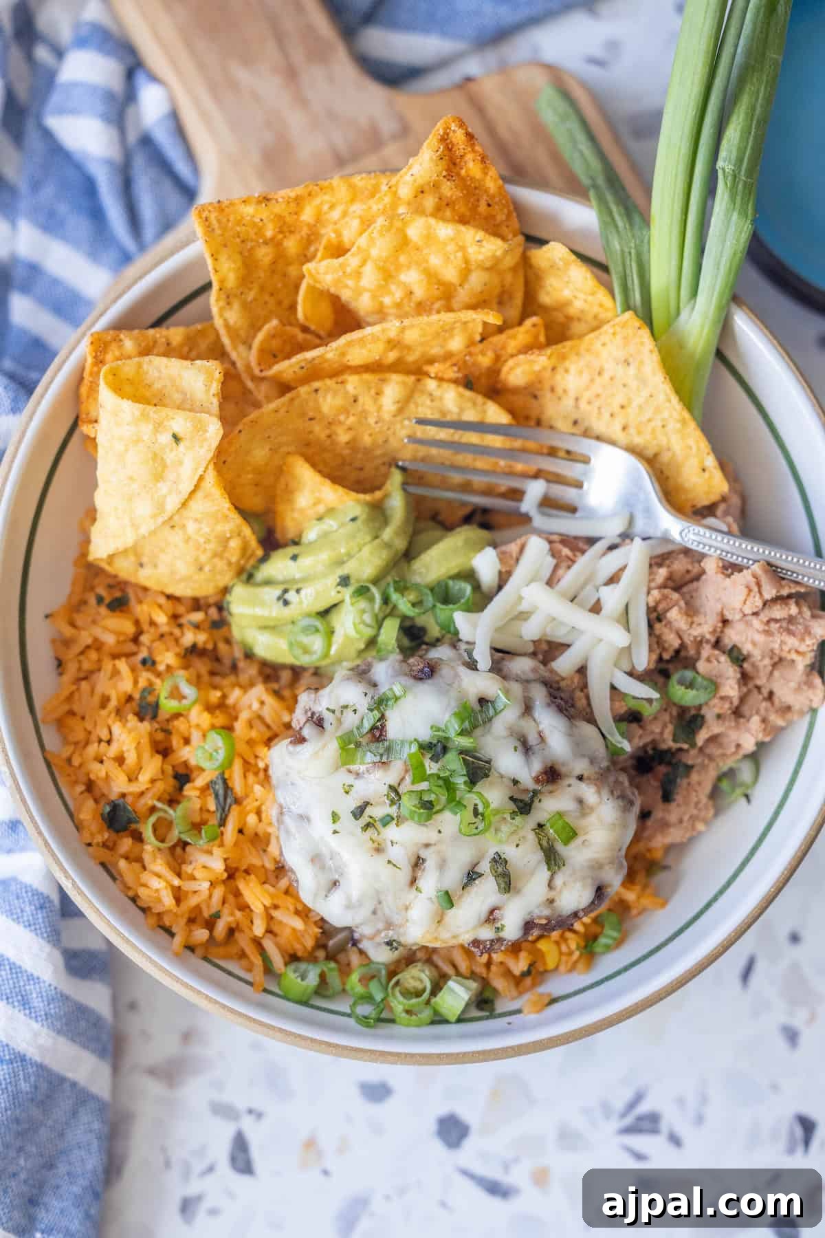 A bowl with Mexican rice, refried beans, a cheesy Green Chile Burger patty, tortilla chips, guacamole, shredded cheese, and green onions, with a fork and extra green onions on the side.