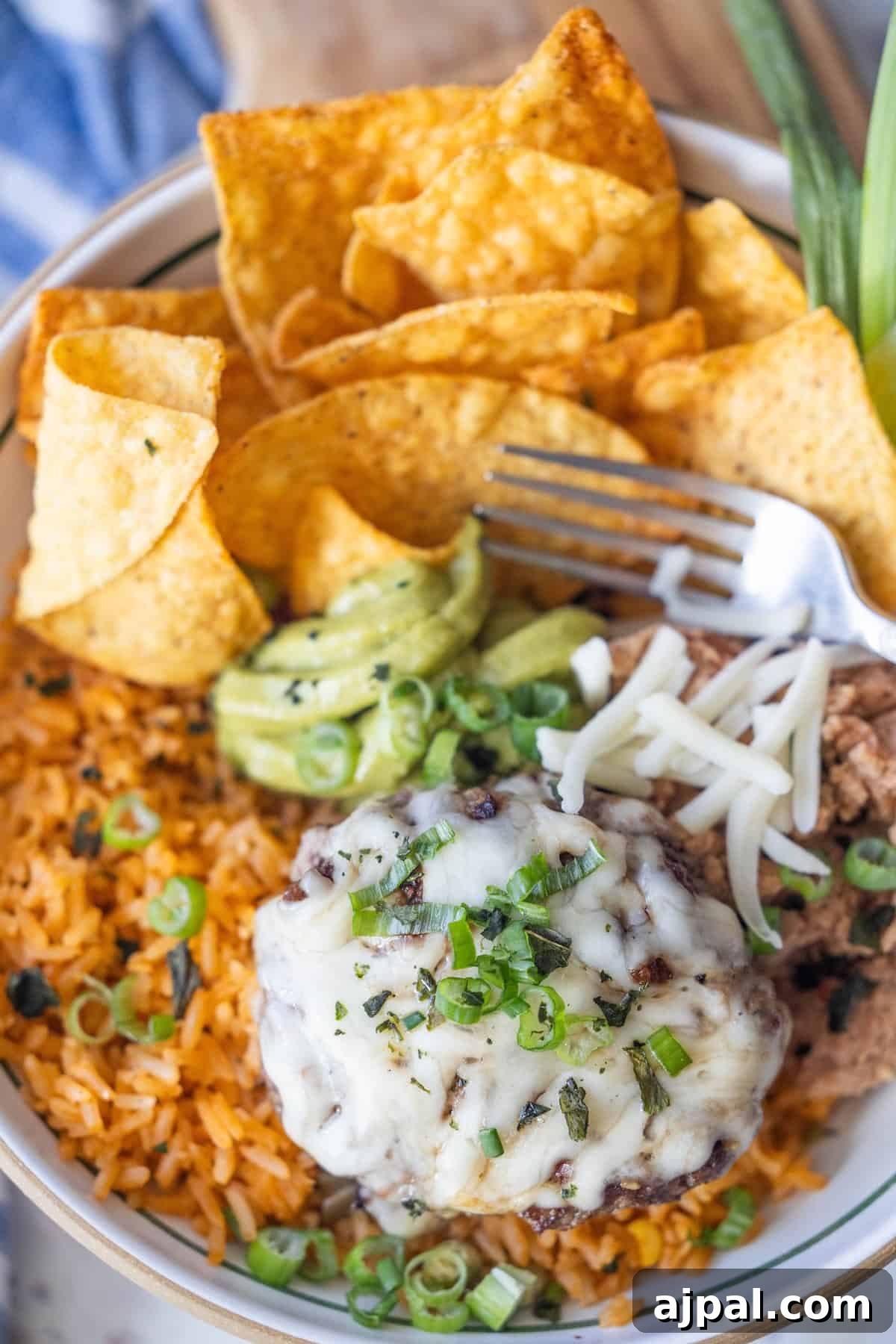 A bowl with Mexican rice, refried beans topped with shredded cheese, a grilled green chile burger patty, avocado slices, tortilla chips, and green onions, with a fork resting on the side.