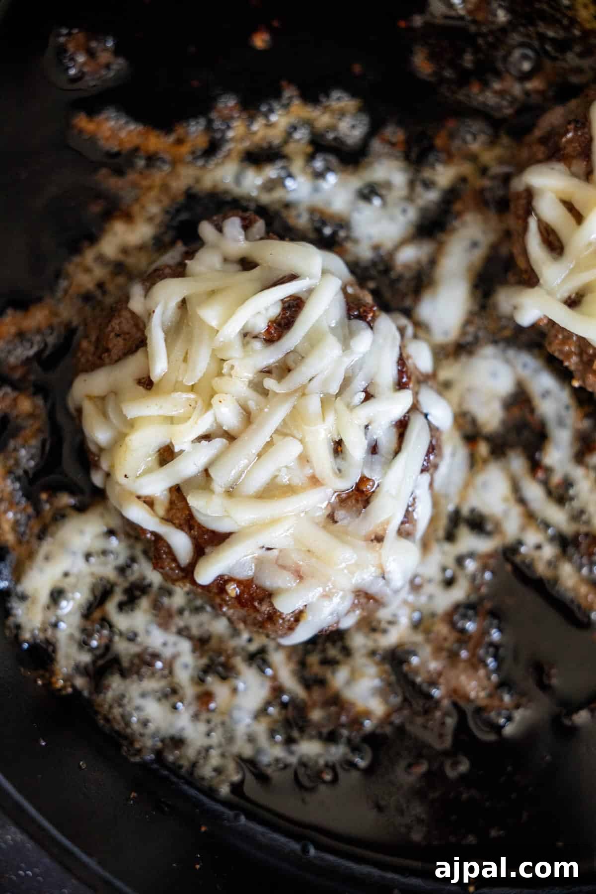 A cooked burger patty in a skillet topped with melted shredded cheese and zesty green chile