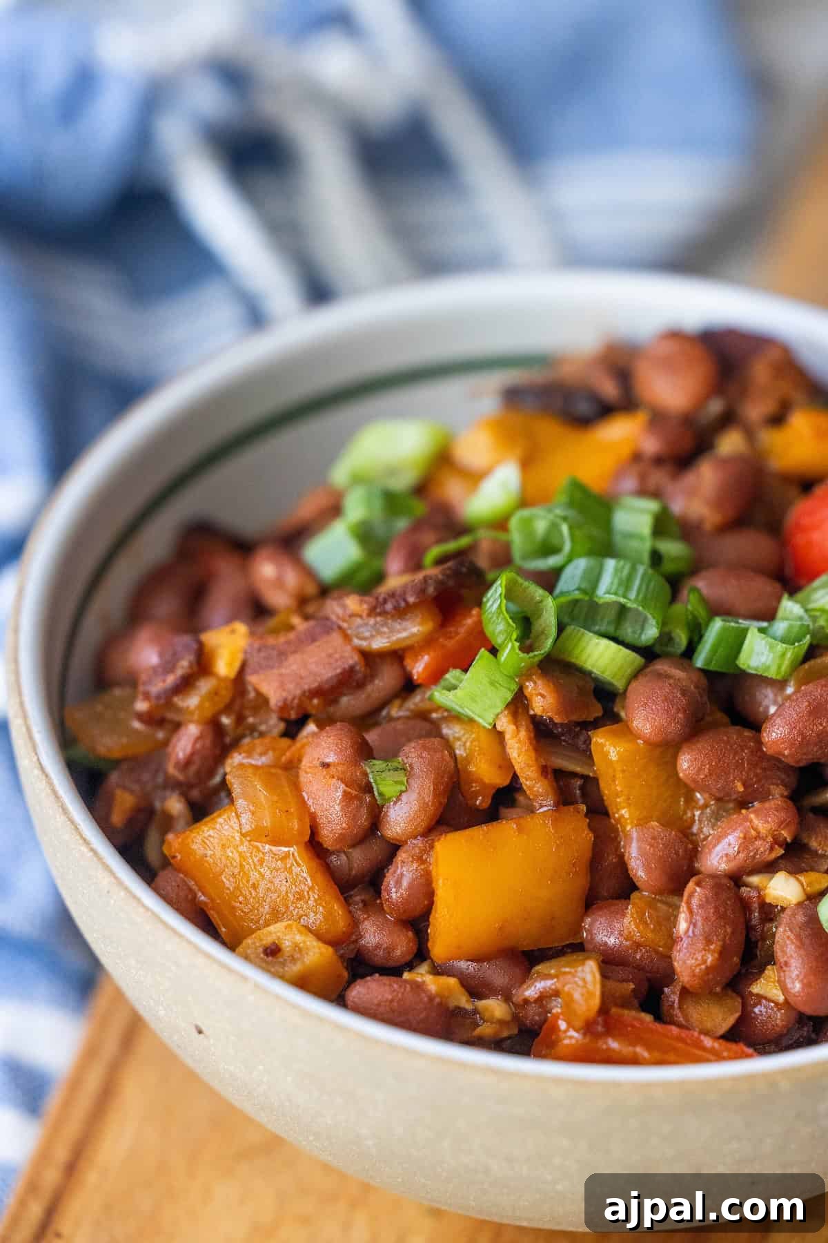 Authentic Mexican Charro Beans Recipe with Bacon and Chiles 3 A bowl of Charro Beans stew with diced yellow bell peppers, tomatoes, and chopped green onions on top, set on a wooden surface with a blue and white cloth in the background—an authentic recipe for Mexican beans lovers.
