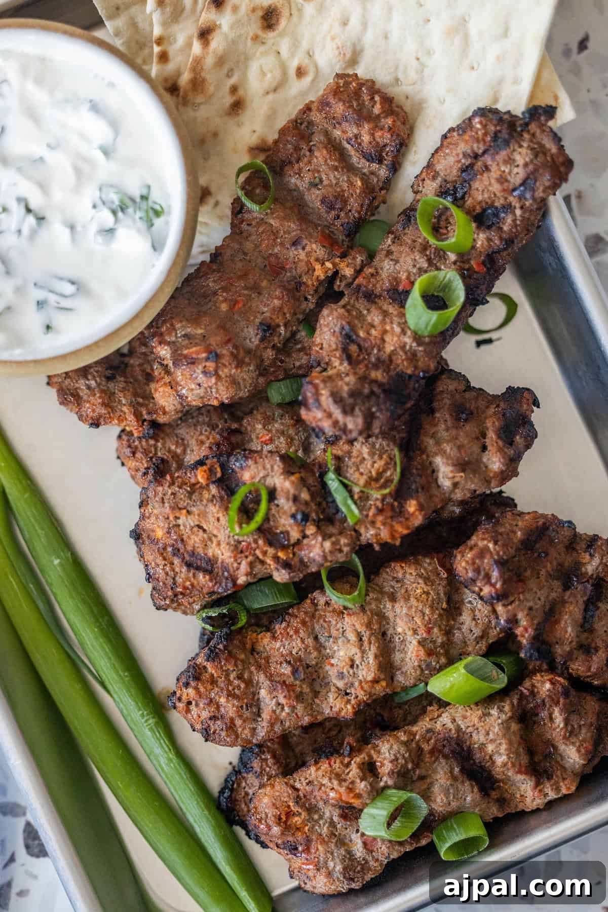 Armenian Spiced Lamb Kebabs Recipe with Authentic Seasoning 2 Armenian spiced meat kebabs garnished with sliced green onions, served with flatbread, extra green onions, and a bowl of yogurt sauce on a metal tray.