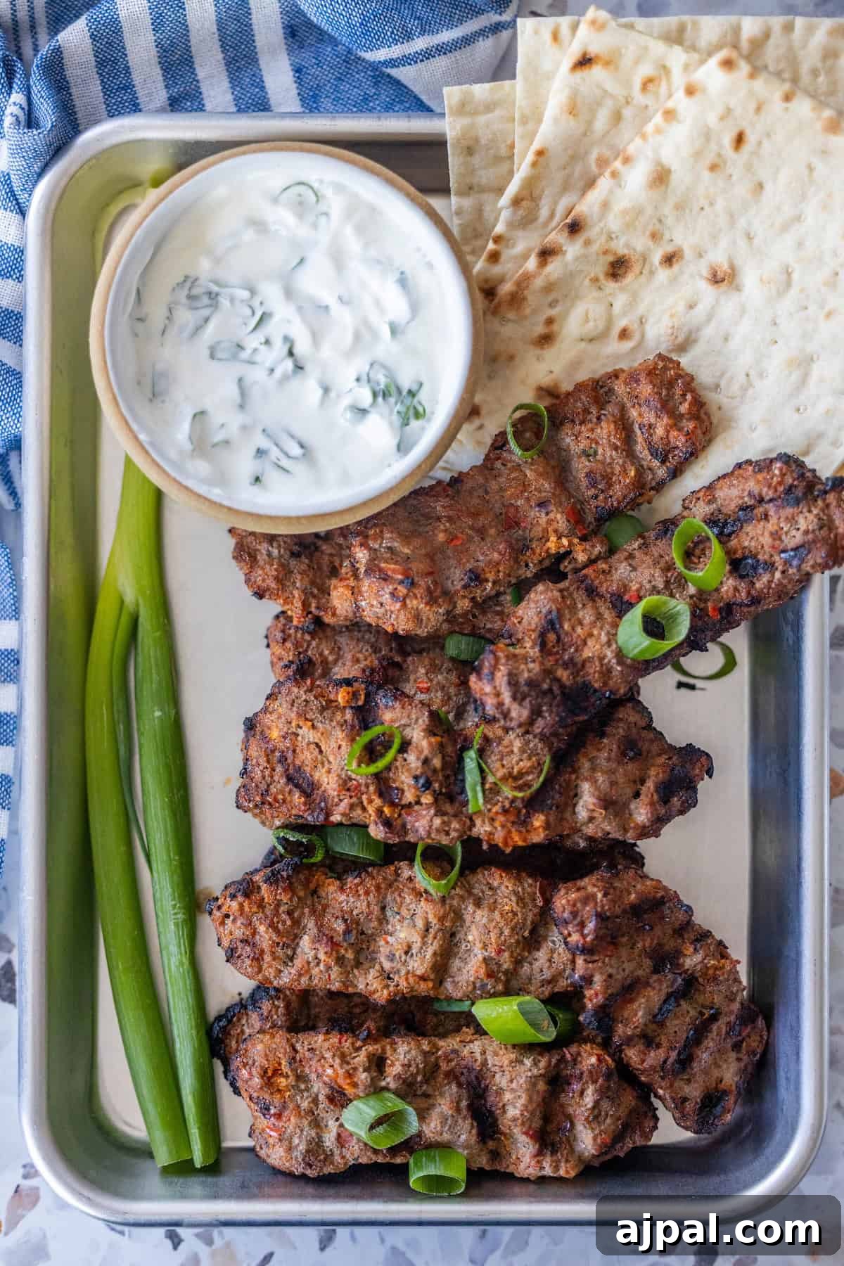 Armenian Spiced Lamb Kebabs Recipe with Authentic Seasoning 11 A metal tray with Armenian spiced meat kebabs, sliced green onions, flatbread, two whole green onions, and a bowl of yogurt sauce.