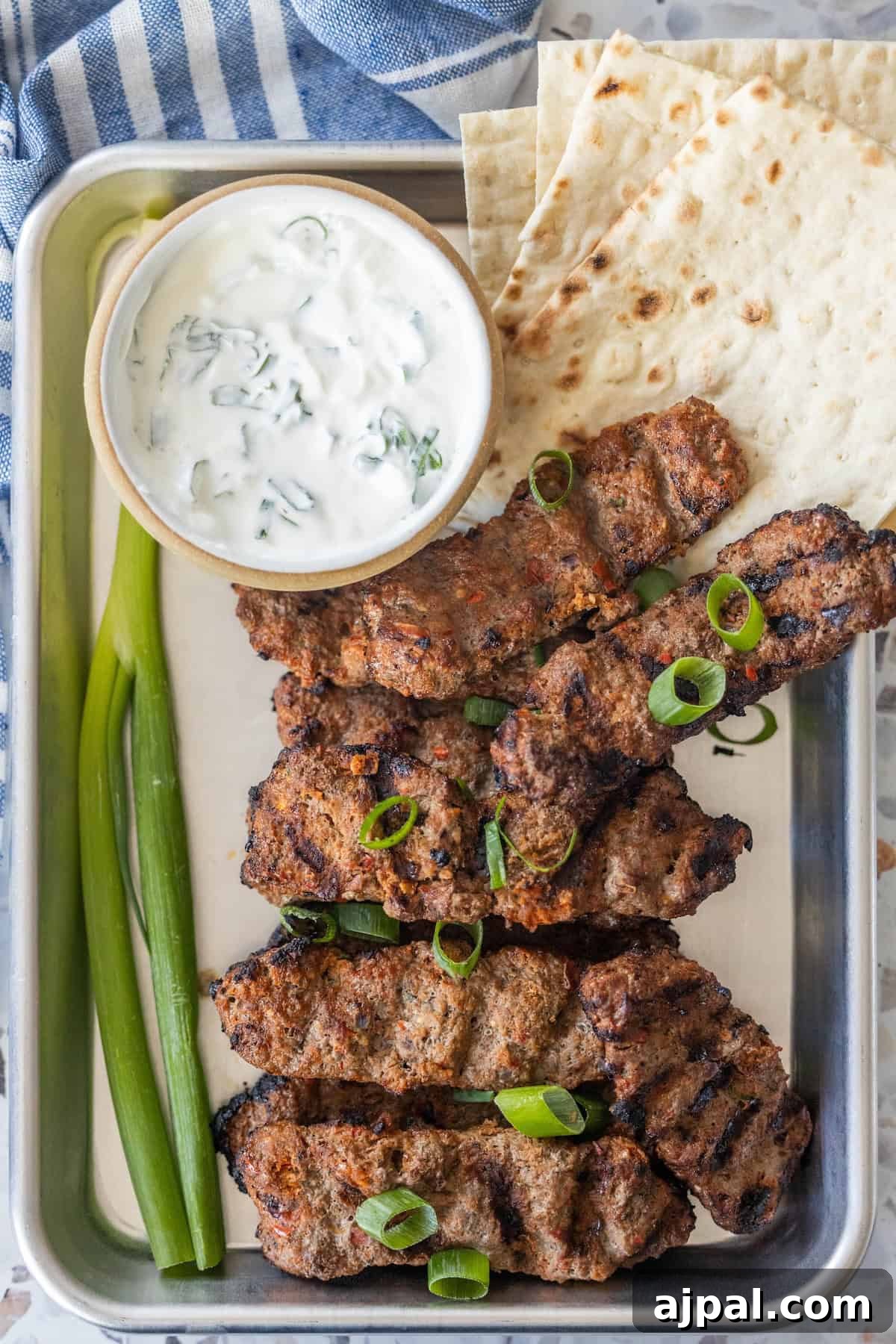 Armenian Spiced Lamb Kebabs Recipe with Authentic Seasoning 4 Tray with Armenian spiced meat kebabs topped with sliced green onions, flatbread, two green onions, and a bowl of yogurt sauce with herbs.