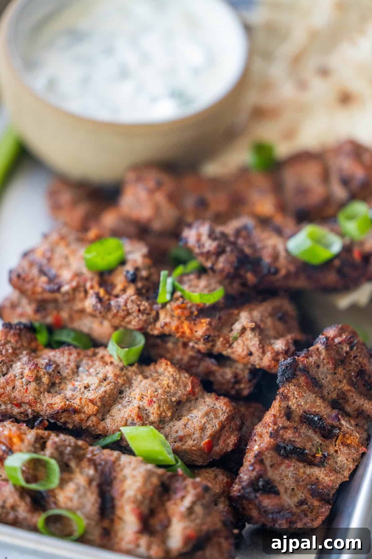 Armenian Spiced Lamb Kebabs Recipe with Authentic Seasoning 5 Armenian spiced meat kebabs garnished with chopped green onions, served with a bowl of creamy dipping sauce and flatbread in the background.