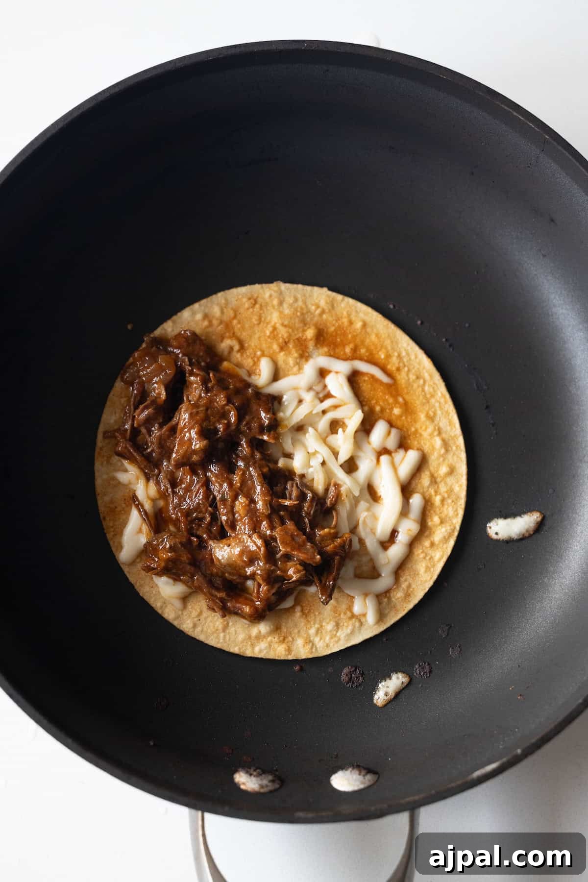 Authentic Birria Tacos Recipe: Juicy Mexican Beef Tacos 11 A corn tortilla in a black skillet topped with shredded meat and grated cheese.