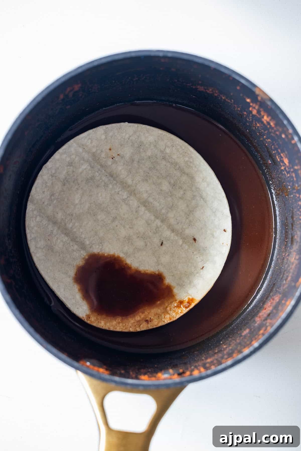 Authentic Birria Tacos Recipe: Juicy Mexican Beef Tacos 10 A corn tortilla is partially submerged in red sauce in a small pot.