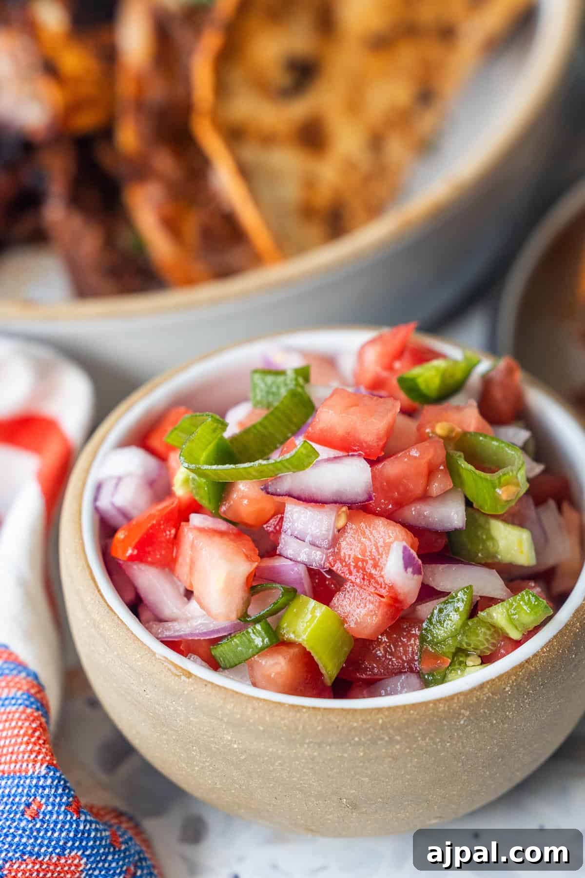 Pico de Gallo Recipe: Fresh Mexican Tomato Salsa 3 A bowl of chopped tomatoes, red onions, and green chili peppers sits on a table next to a plate of food and a colorful napkin.