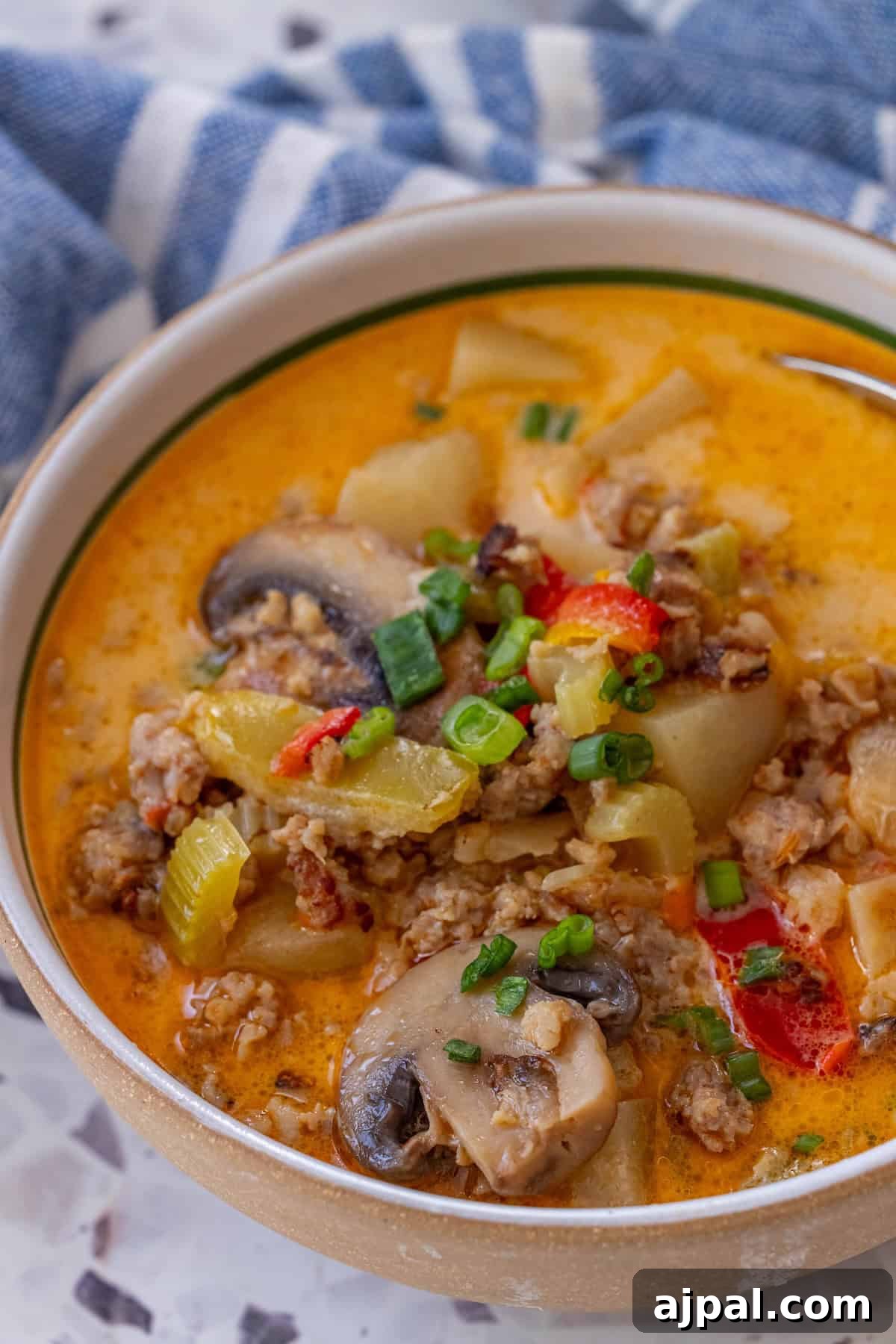 30 Back-to-School Meals That Reheat Perfectly 13 A bowl of 20 Minute Creamy Sausage and Mushroom Soup with sliced mushrooms, ground meat, potatoes, veggies, and green onions sits on a marble surface near a blue and white cloth.