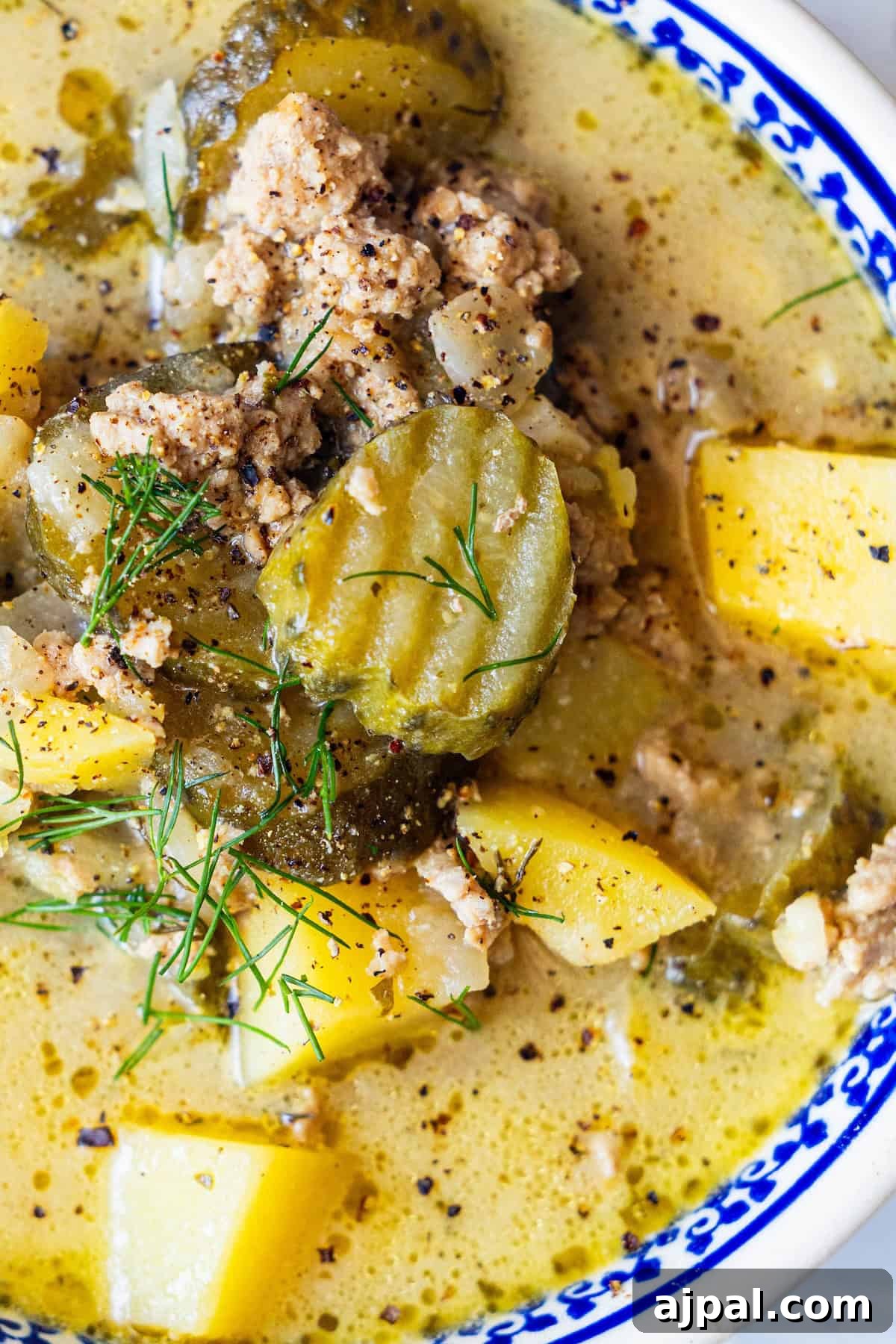 Close-up of creamy soup with ground meat, sliced pickles, potatoes, and cracked black pepper.