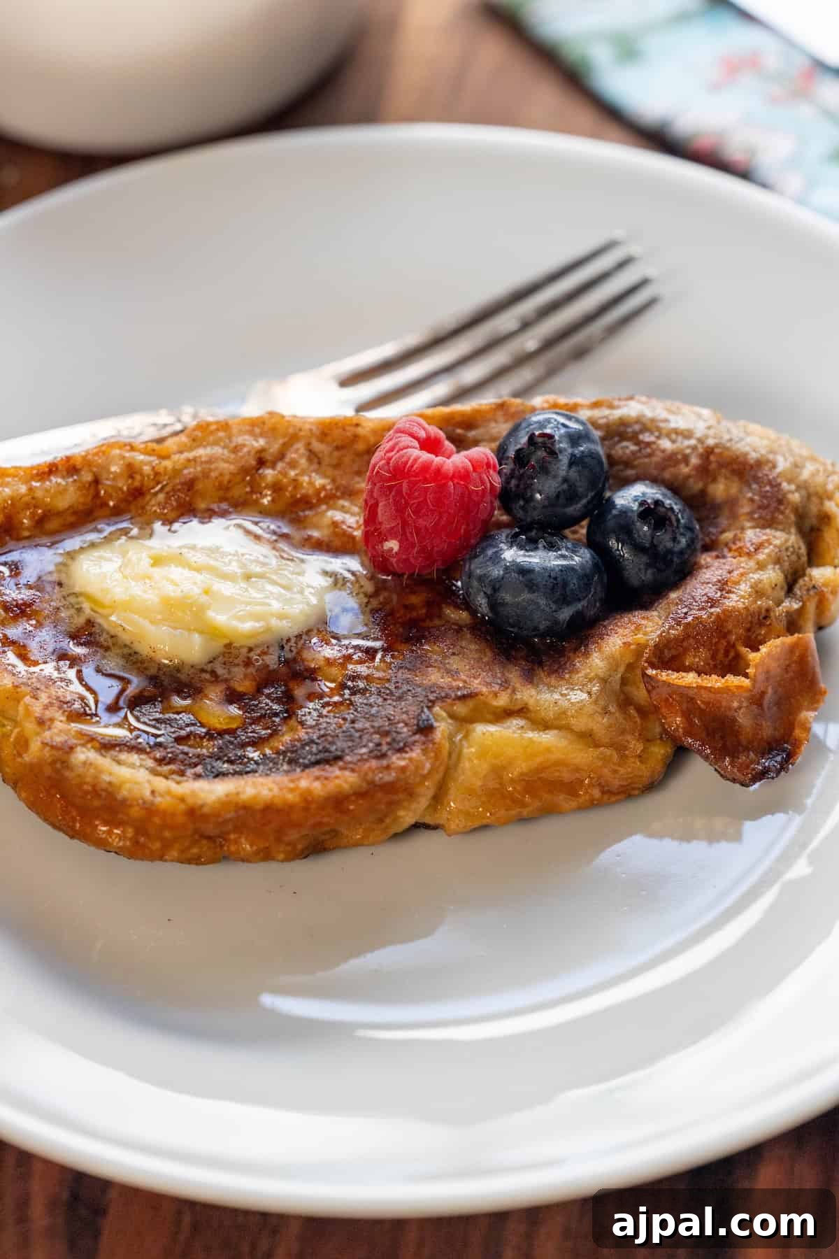 A slice of Challah French Toast topped with butter, syrup, a raspberry, and two blueberries rests on a white plate with a fork.