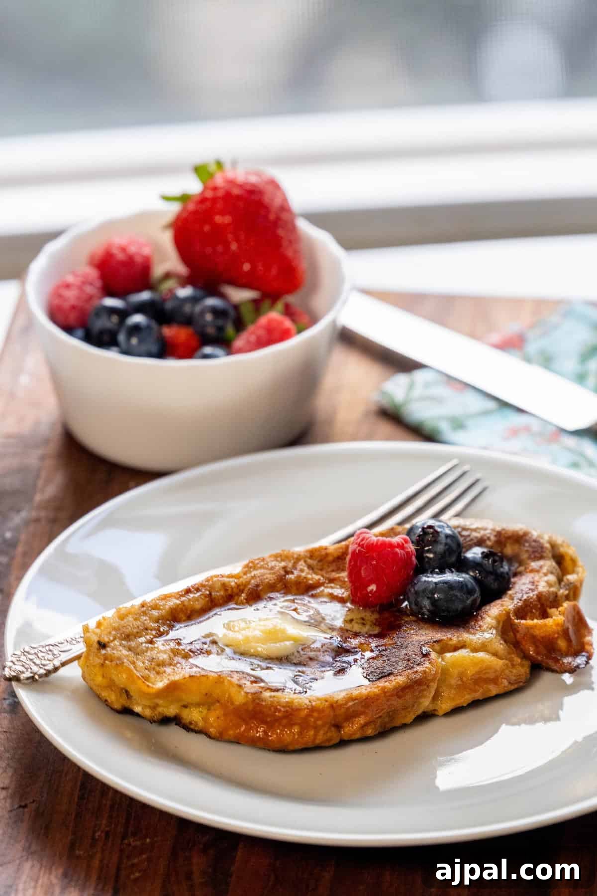 A slice of cinnamon sugar French toast made with rich challah bread sits on a white plate, topped with butter, blueberries, and a raspberry, alongside a fork; a bowl of mixed berries is in the background on a wooden table.