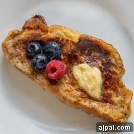 A slice of rich Challah French Toast is topped with a pat of butter, three blueberries, and one raspberry on a white plate.