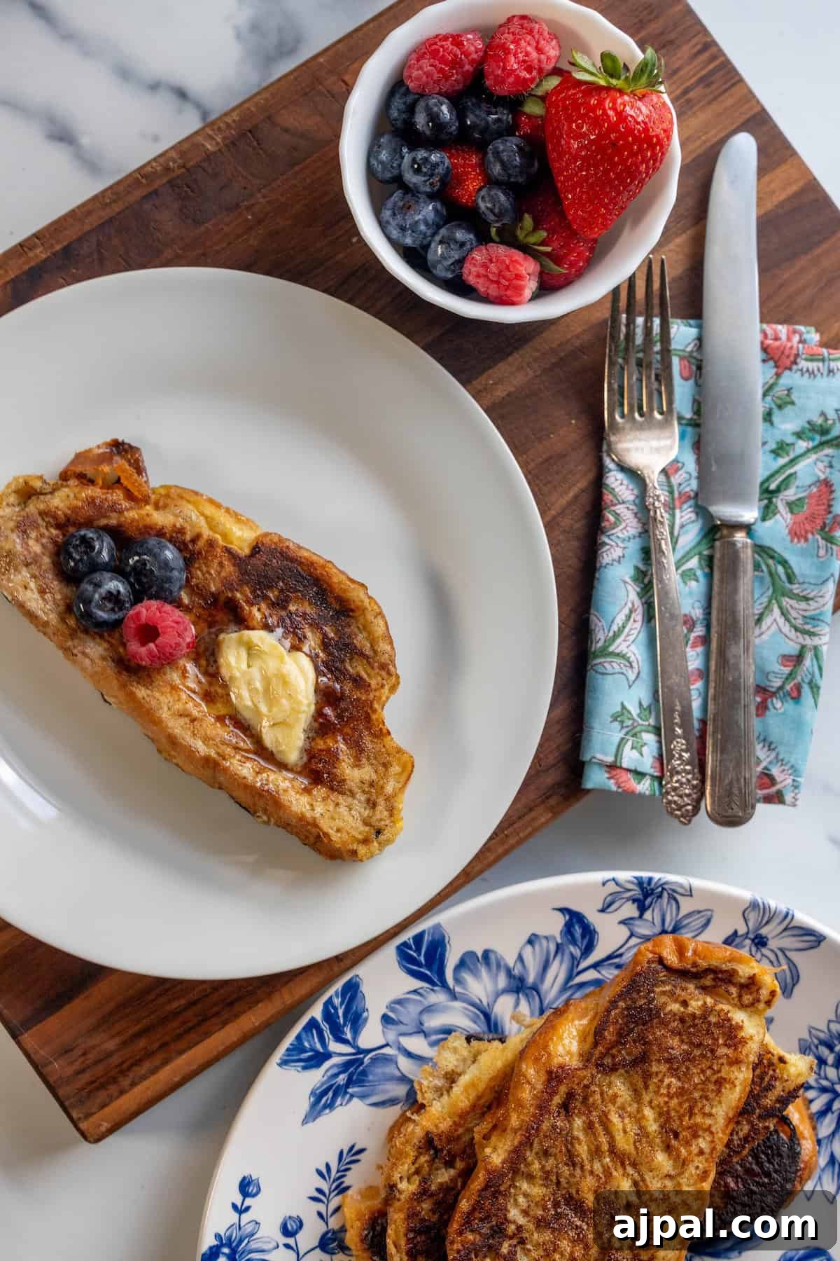 A plate with rich Challah French Toast topped with butter and berries, a bowl of mixed berries, utensils on a napkin, and another plate with more French Toast on a wooden board.