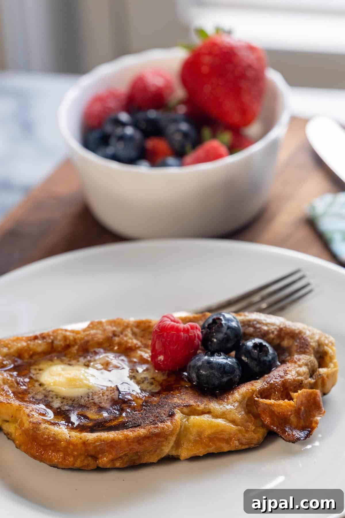 A slice of indulgent Challah French Toast topped with butter, blueberries, and a raspberry on a white plate, with a fork and a bowl of mixed berries in the background.