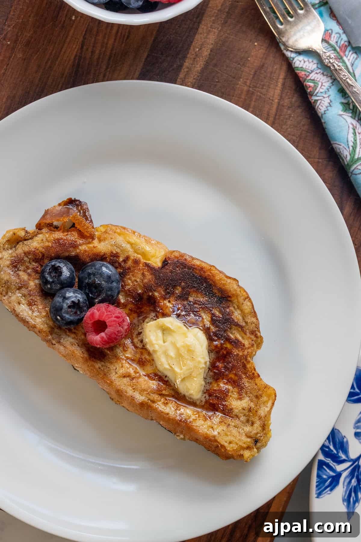 A slice of Challah French Toast with butter, blueberries, and a raspberry on a white plate, set on a wooden surface next to a fork and a small bowl of berries.