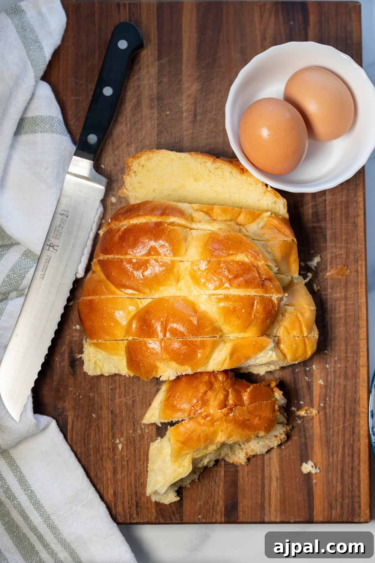 A loaf of braided challah bread is sliced on a wooden cutting board with a serrated knife, next to a white bowl holding two brown eggs and a striped kitchen towel—perfect for making cinnamon sugar French toast.