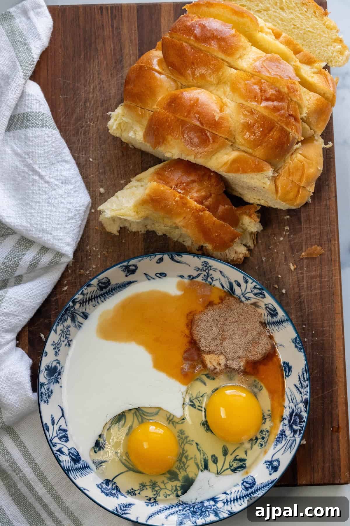 Sliced braided challah bread on a wooden board next to a bowl with two eggs, milk, sugar, and vanilla extract—perfect ingredients for making French Toast. A white-striped towel is on the side.