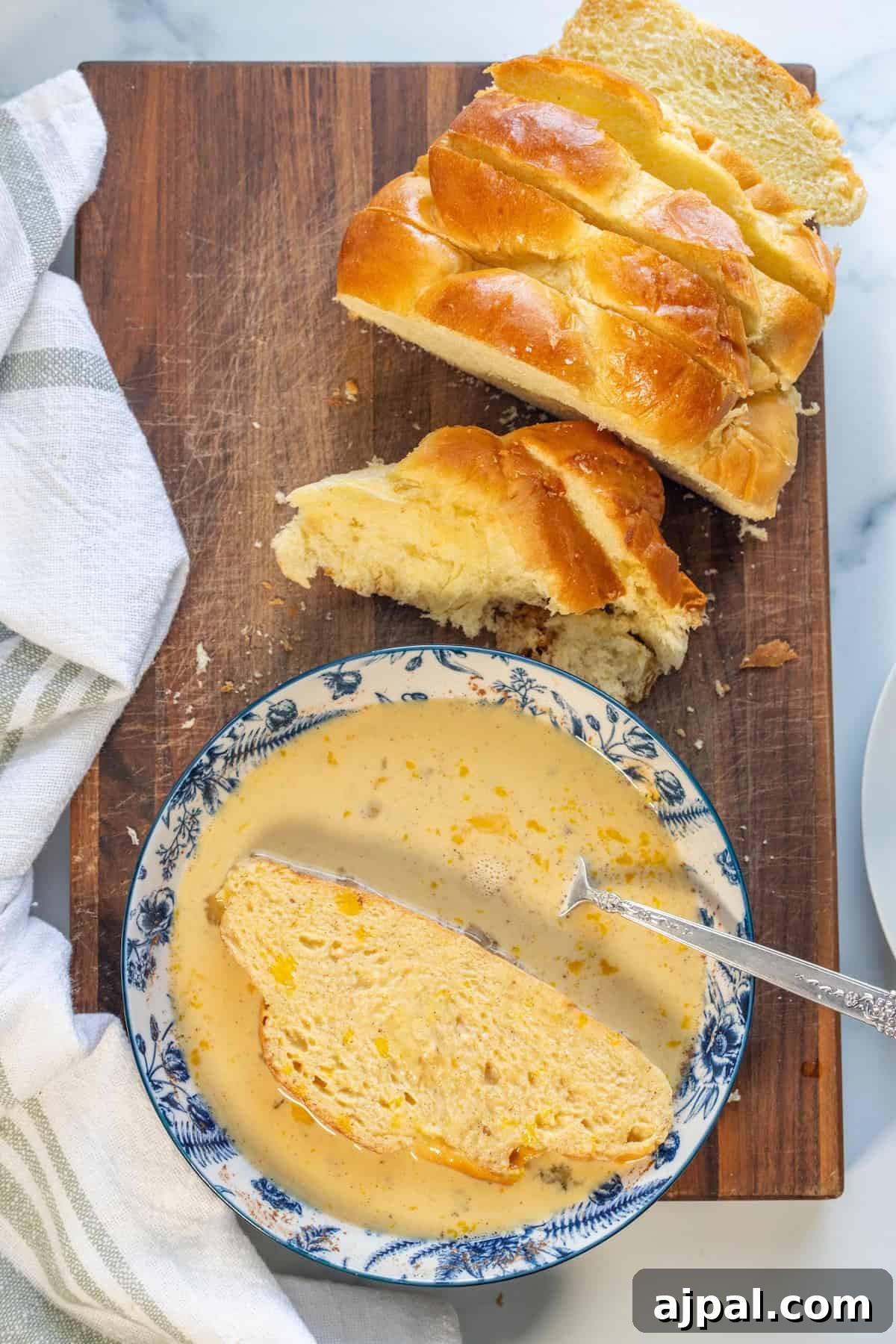 A slice of braided bread in a bowl of soup with a spoon, next to more sliced bread on a wooden cutting board with a white towel nearby—perfect for transforming into leftover challah French toast the next morning.
