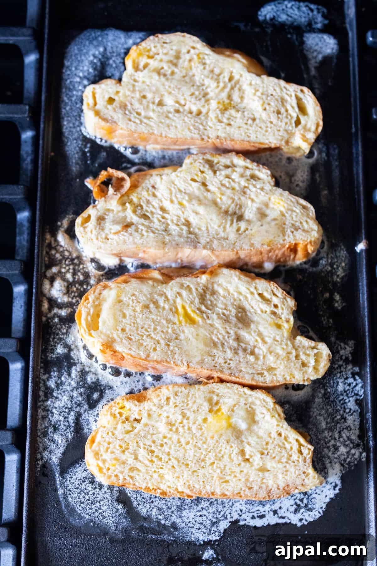 Four slices of bread coated in egg mixture are cooking on a buttered griddle, making classic French toast with rich Challah bread and a hint of cinnamon sugar.