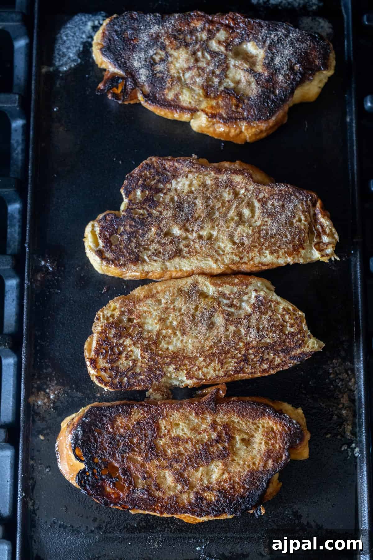 Four slices of French toast are cooking on a griddle, made from thick-cut Challah and topped with cinnamon sugar, with visible browning and crispy edges on each piece.